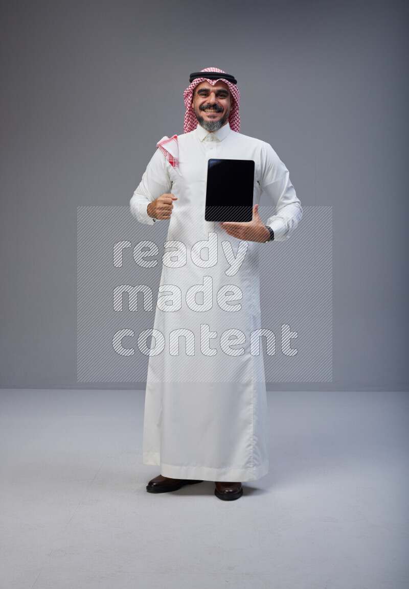 Saudi man Wearing Thob and red Shomag standing showing tablet to camera on Gray background