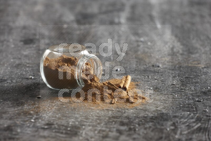 Flipped glass herbs jar full of cinnamon powder with cracked cinnamon sticks on a textured black background