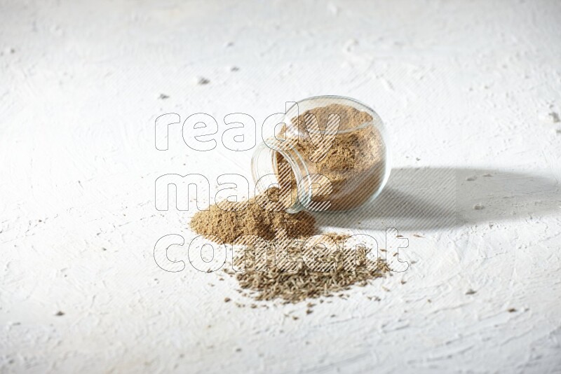 A flipped glass spice jar full of cumin powder and powder spilled out with cumin seeds on textured white flooring