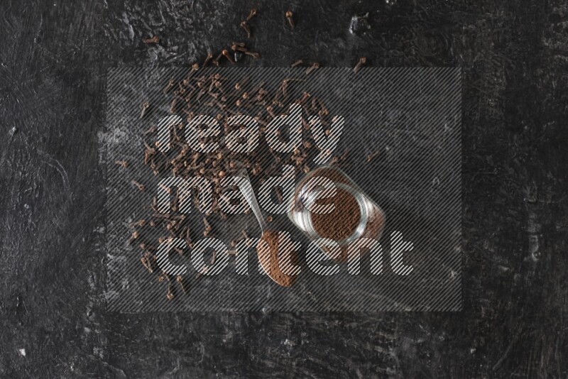 A glass spice jar and a metal spoon full of cloves powder with cloves spread on textured black flooring