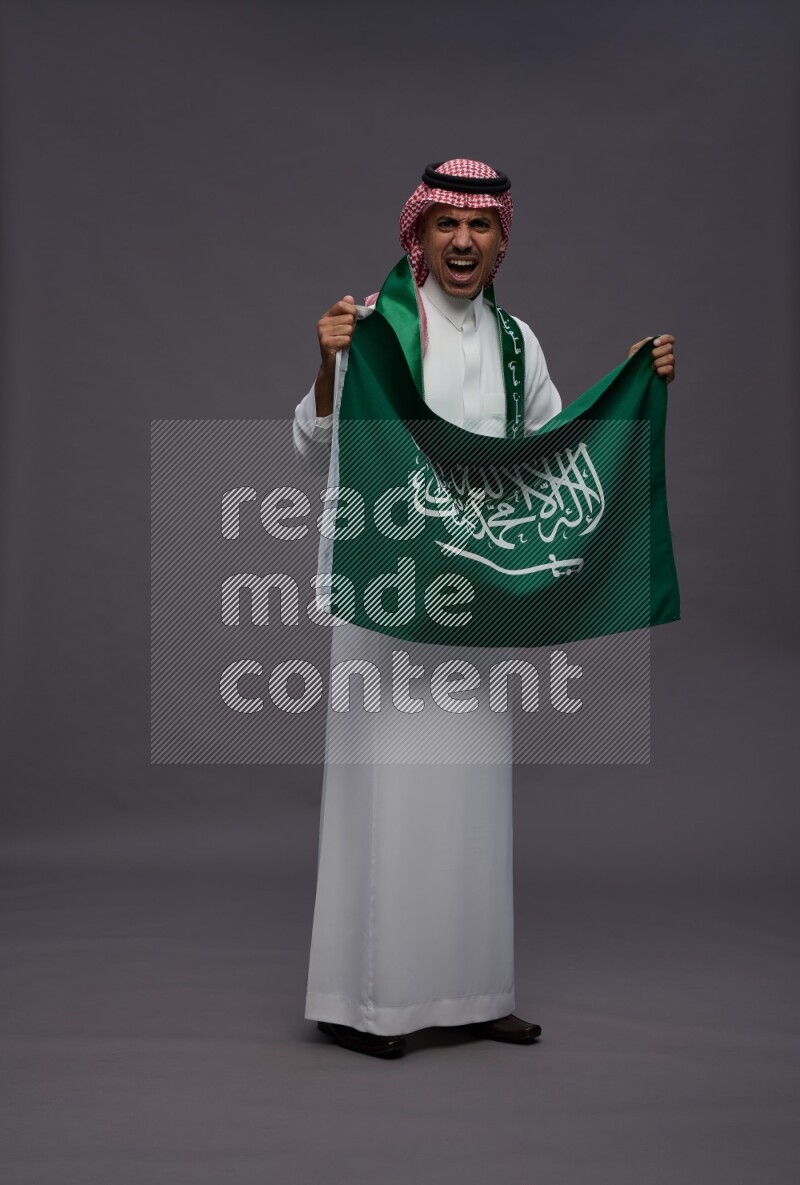 Saudi man wearing thob and shomag standing holding Saudi flag on gray background