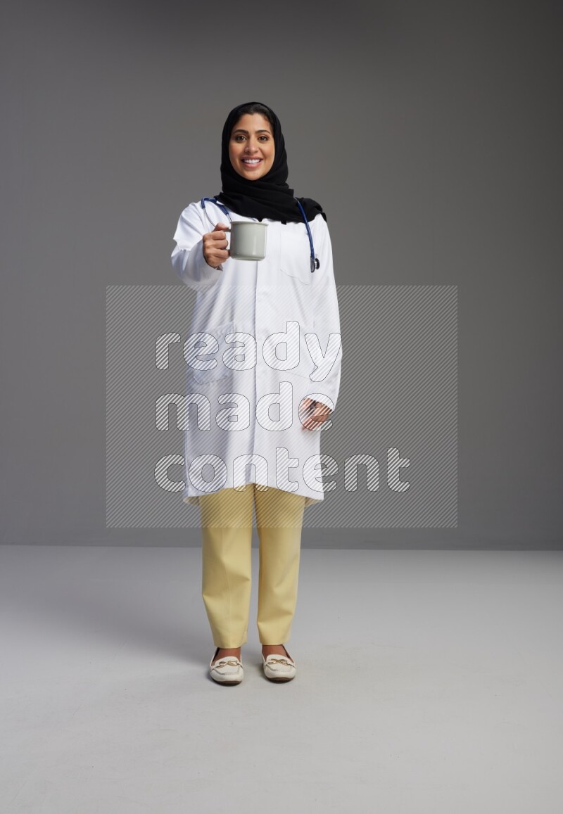 Saudi woman wearing lab coat with stethoscope standing holding mug on Gray background