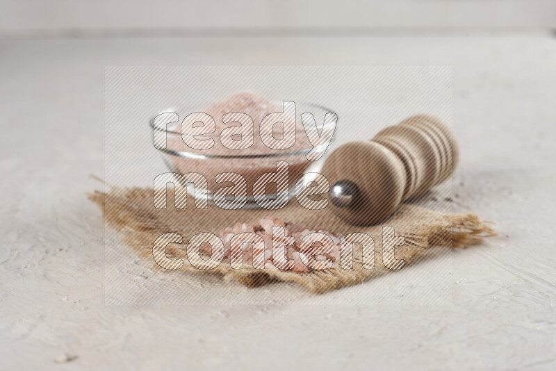 A glass bowl full of pink himalayan salt with a wooden grinder on a burlap fabric all on white background