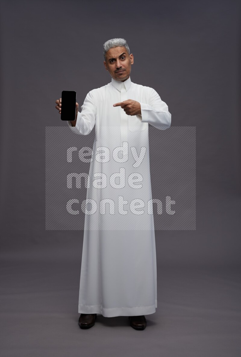 Saudi man wearing thob standing showing phone to camera on gray background