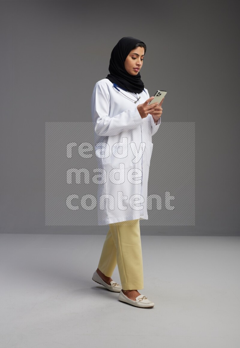 Saudi woman wearing lab coat with stethoscope standing texting on phone on Gray background
