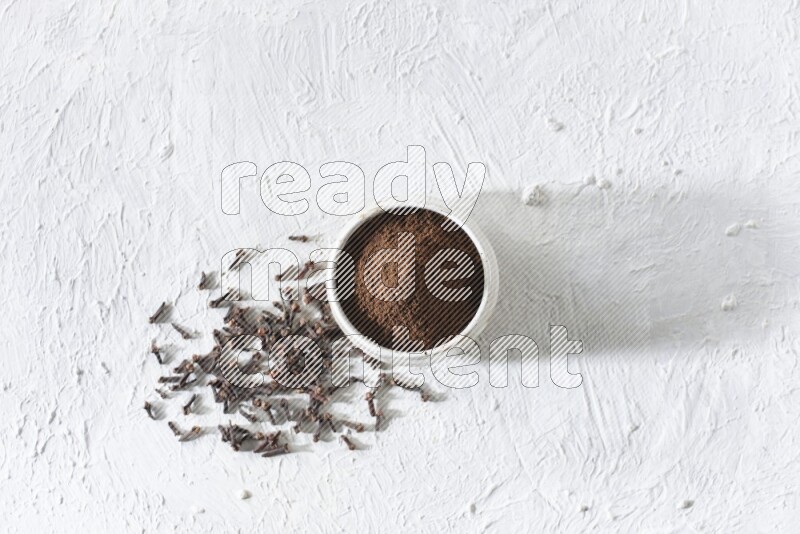 وعاء بيج من السيراميك ممتلئ ببودرة القرنفل وحبوب القرنفل على أرضية بيضاء