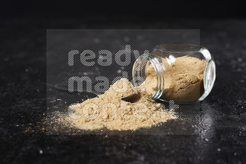 A glass jar full of ground ginger powder flipped with some spilling powder on black background
