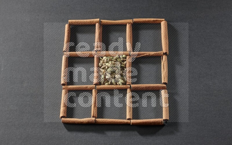 9 squares of cinnamon sticks full of cardamom in the middle surrounded by nutmeg, cinnamon, bay laurel leaves, cloves, cumin, dried ginger, dried basil and star anise on black flooring