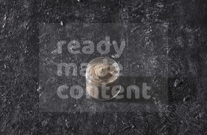 A glass jar full of black pepper powder on a textured black flooring