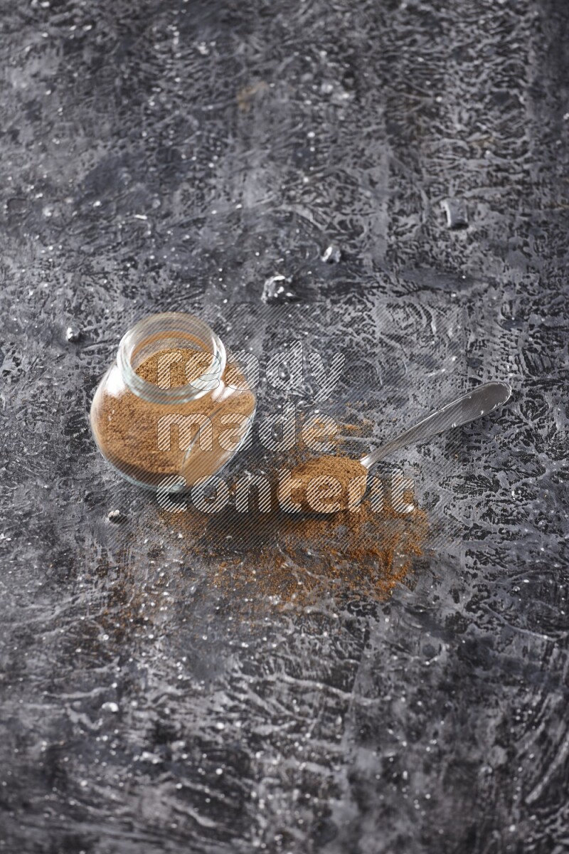 Herbal glass jar full of cinnamon powder and a metal spoon full of powder on textured black background