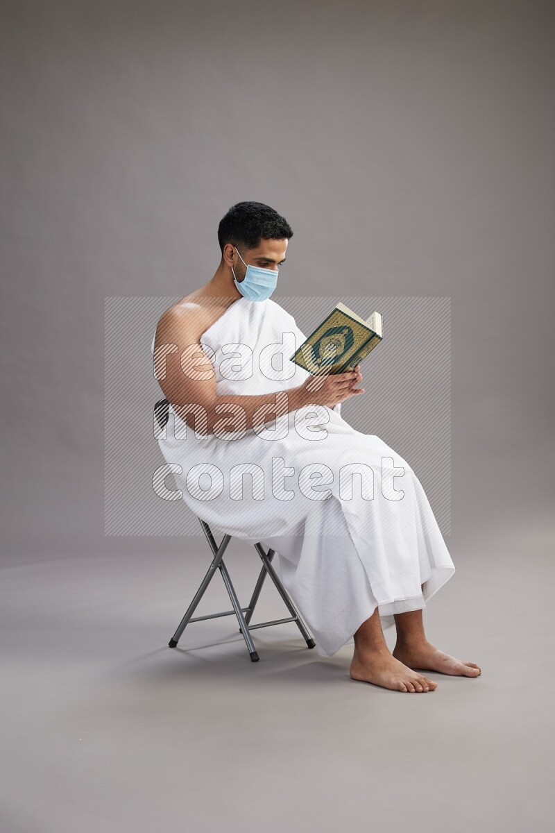 A man wearing Ehram with face mask sitting on chair reading quran on gray background