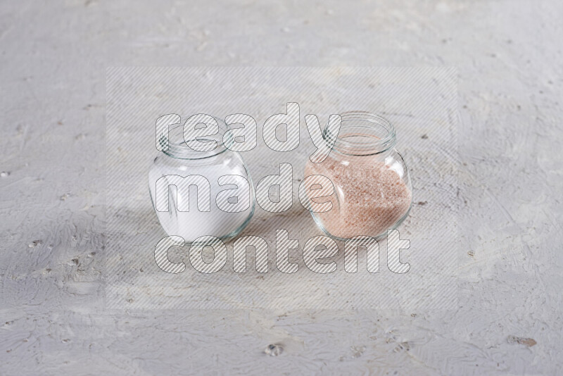 2 glass jars one is full of fine himalayan salt and the other with table salt on white background