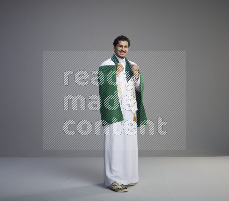 A Saudi man standing wearing thob with face painting wrapping Saudi flag scarf and holding big Saudi flag on gray background