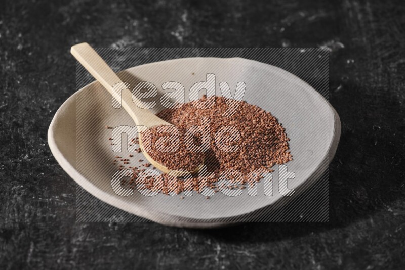 A multicolored pottery plate full of garden cress seeds and wooden spoon full of seeds on a textured black flooring