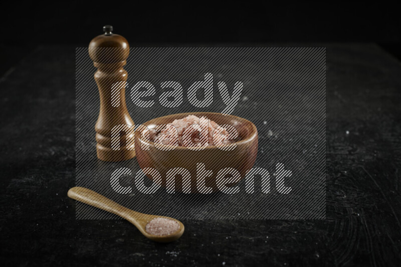 A wooden bowl and spoon filled with coarse pink himalayan salt and a wooden grinder beside them on black background
