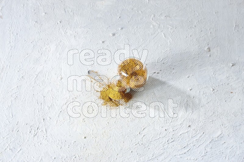 وعاء زجاجي للتوابل مقلوب وملعقة معدنية ممتلئان ببودرة الكركم والبودرة تناثرت منه على خلفية بيضاء