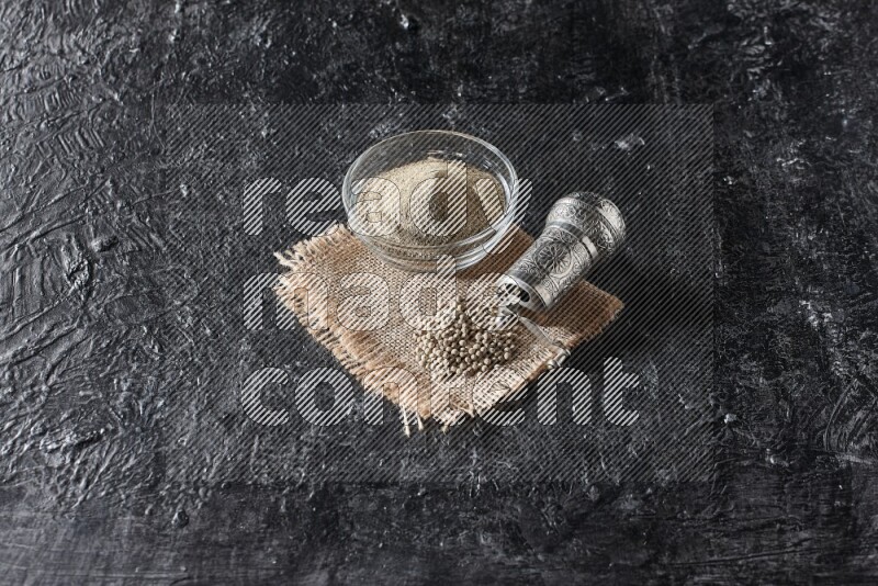 A glass bowl full of white pepper powder with white pepper beads on a burlap piece of fabric and a metal grinder on textured black flooring