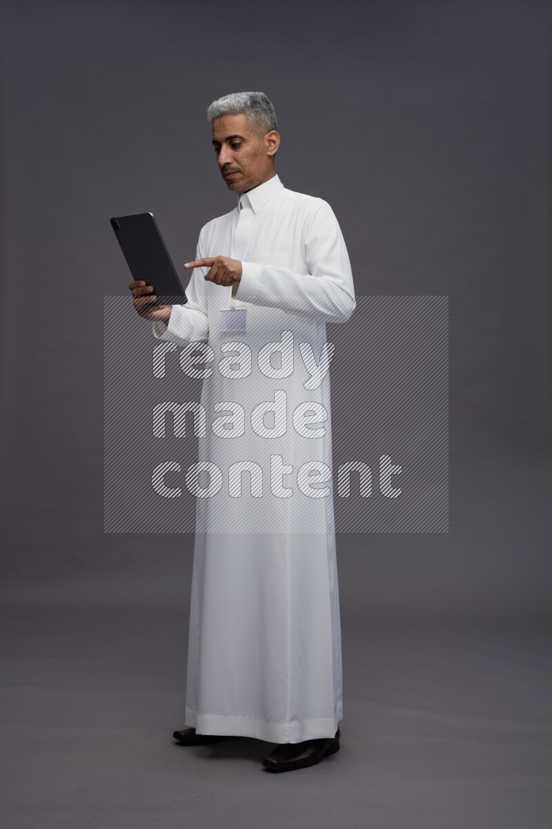 Saudi man wearing thob with neck strap employee badge standing working on tablet on gray background
