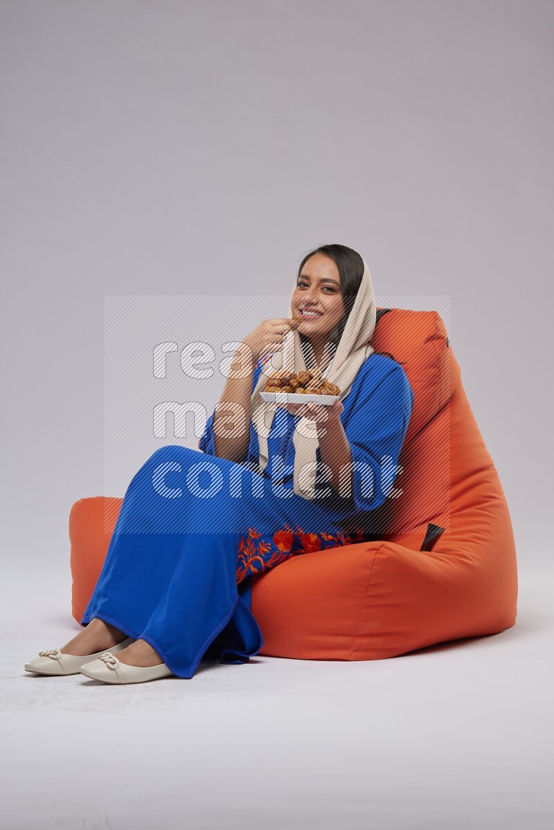 A Woman sitting on an orange beanbag wearing Jalabeya holding a plate of dates