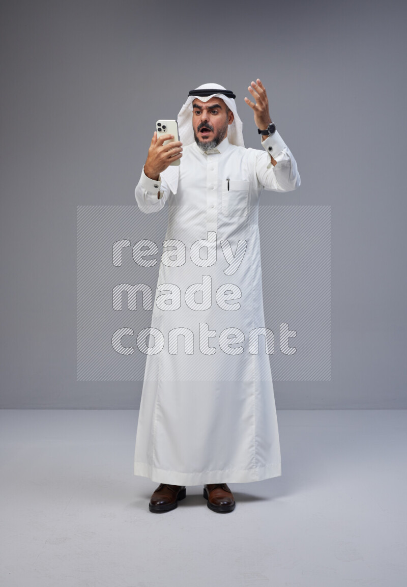 Saudi man Wearing Thob and white Shomag standing texting on phone on Gray background