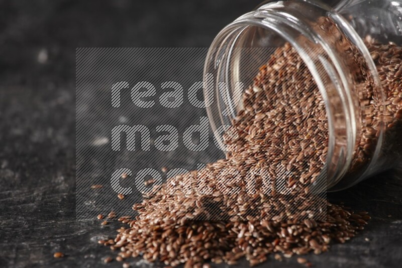 A glass jar full of flaxseeds flipped and seeds spread out on a textured black flooring