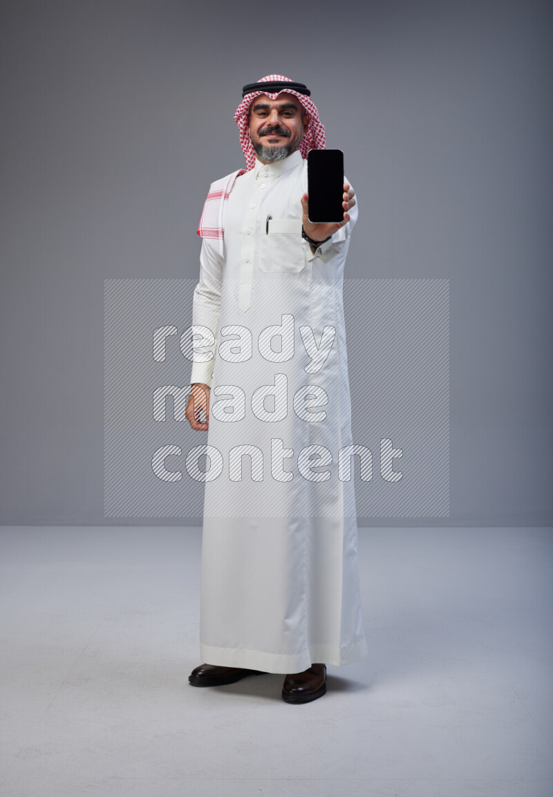 Saudi man Wearing Thob and red Shomag standing showing phone to camera on Gray background