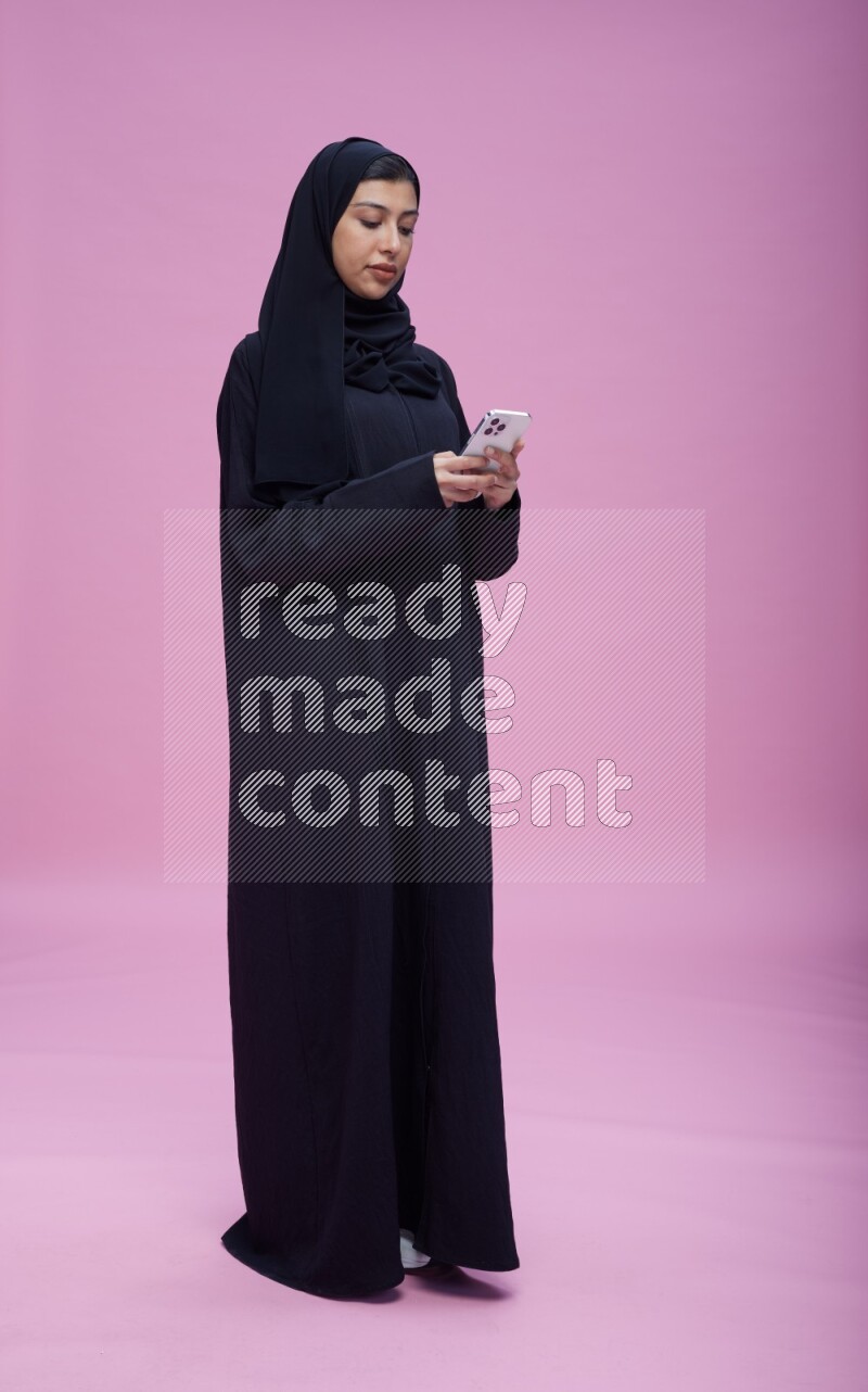 Saudi woman wearing Abaya standing texting on phone on pink background