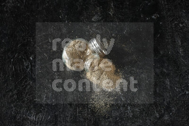 A flipped glass spice jar full of cardamom powder on textured black flooring