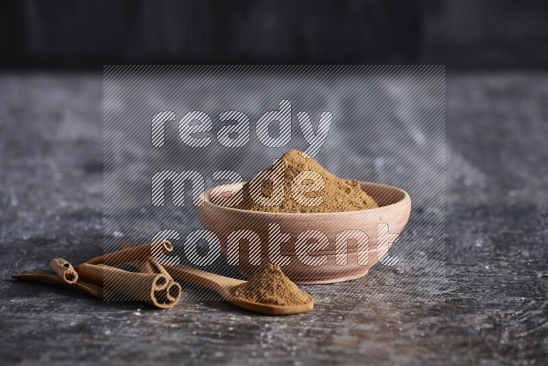 wooden bowl full of cinnamon powder and a wooden spoon full of it with cinnamon sticks on a textured black background