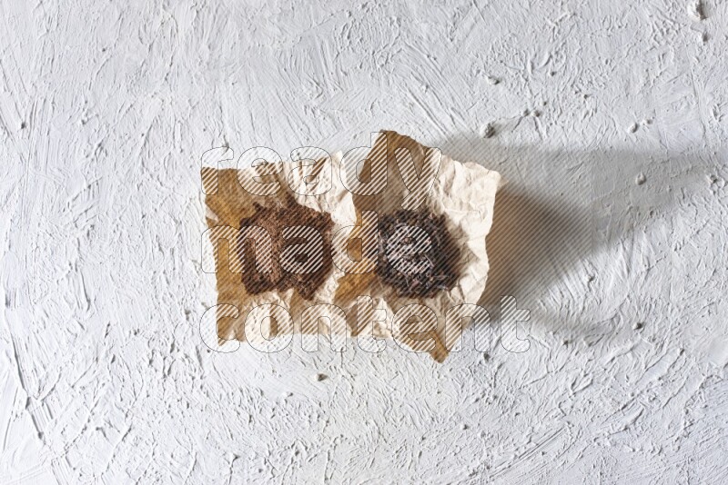 Cloves powder and cloves on 2 crumpled pieces of paper on a textured white flooring