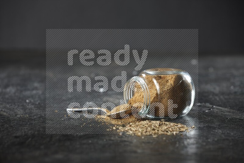 A flipped glass spice jar and a metal spoon full of cumin powder and powder spilled out with cumin seeds on a textured black flooring