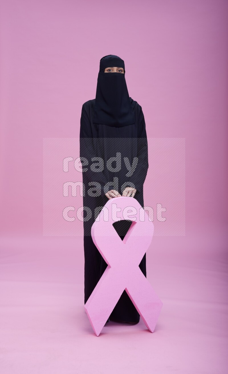 Saudi woman wearing abaya and niqab standing holding awareness ribbon on pink background