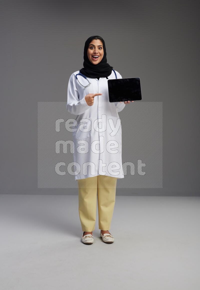 Saudi woman wearing lab coat with stethoscope standing showing tablet to camera with sign in the back on Gray background