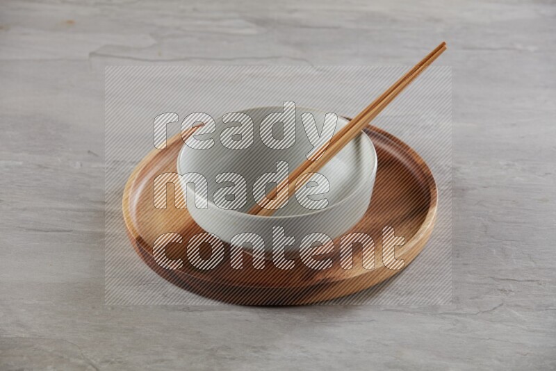 white pottery round bowl on top of brown wood round plate and wood chopsticks, on grey textured countertop