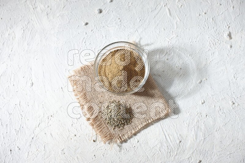 A glass bowl full of cumin powder with some of cumin seeds on burlap piece on a textured white flooring