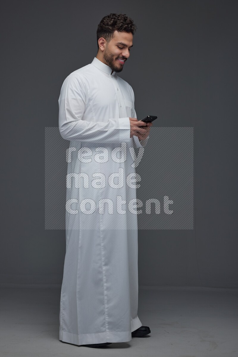 A Saudi man wearing Thobe standing and using his phone eye level on a gray background