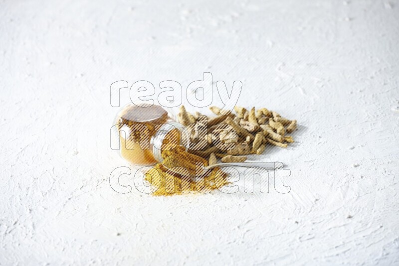 A flipped glass spice jar and metal spoon full of turmeric powder and powder spilled out of it with dried whole fingers on textured white flooring