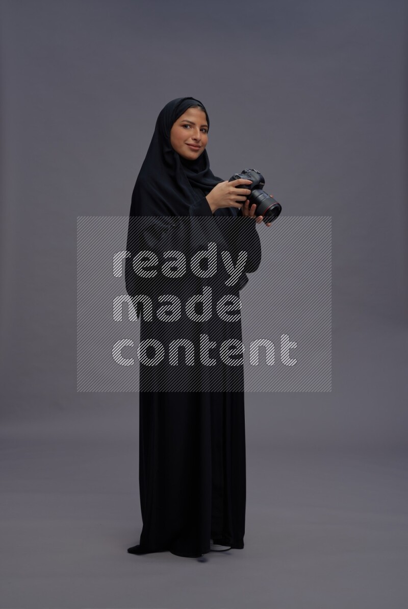 Saudi woman wearing Abaya standing holding Camera on gray background