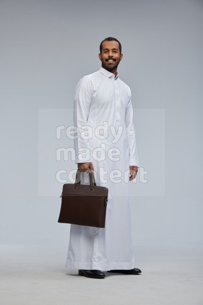 Saudi man wearing thob standing holding suitcase on gray background