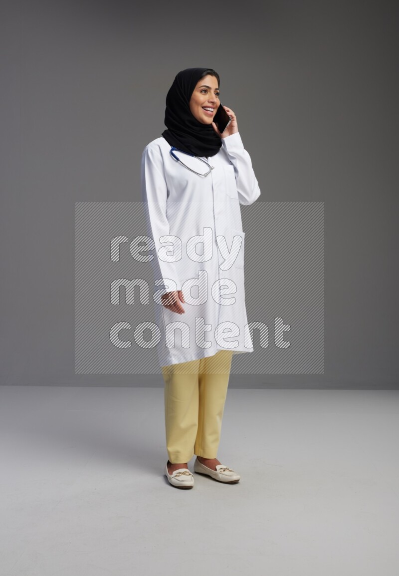 Saudi woman wearing lab coat with stethoscope standing talking on phone on Gray background