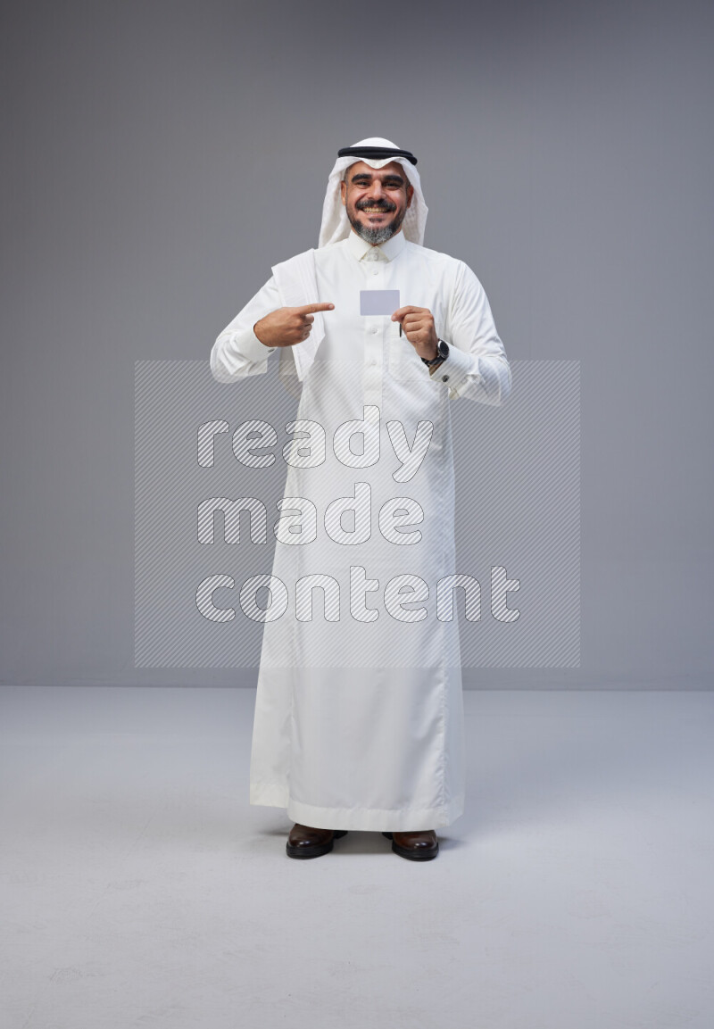 Saudi man Wearing Thob and white Shomag standing holding ATM card on Gray background