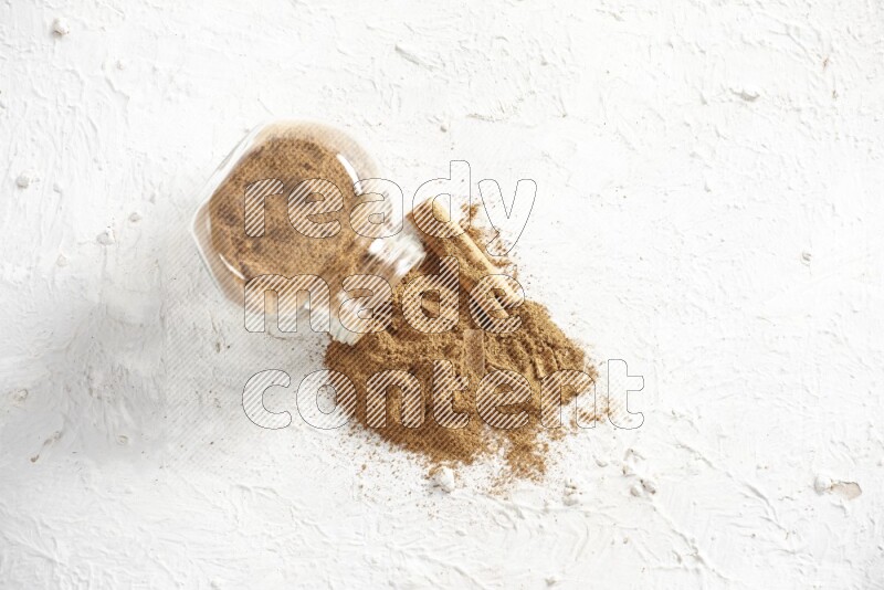 Flipped glass jar full of cinnamon powder with some pieces of cinnamon sticks on a textured white background