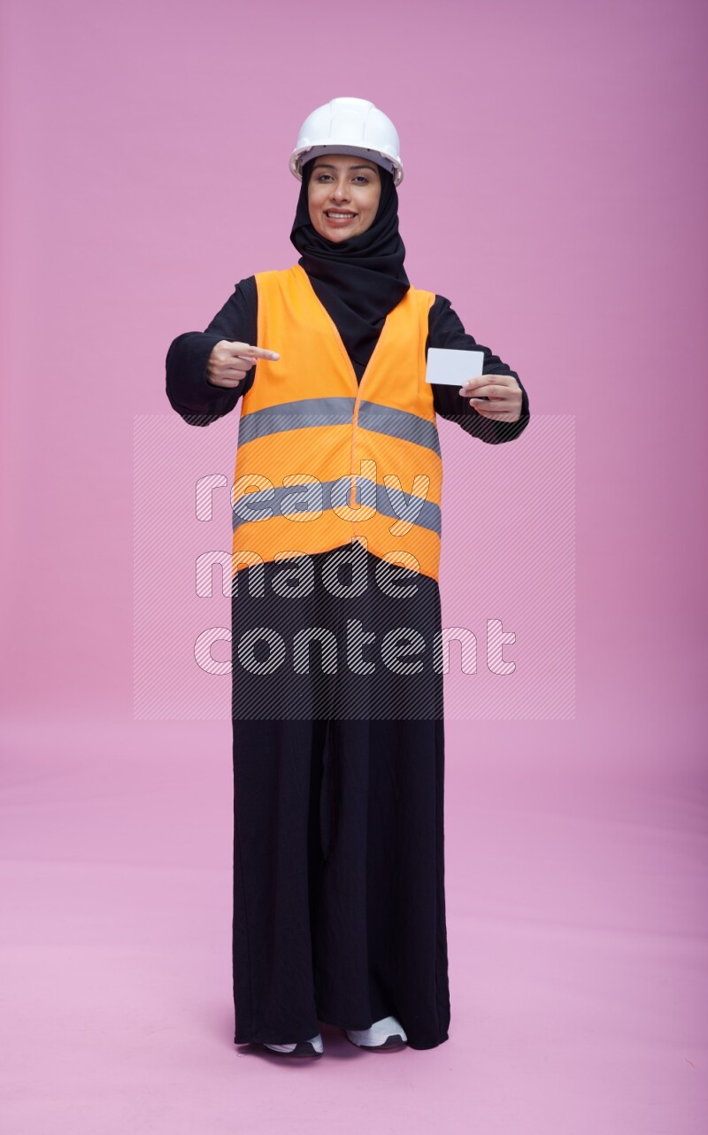 Saudi woman wearing Abaya with engineer vest and helmet standing holding ATM card on pink background