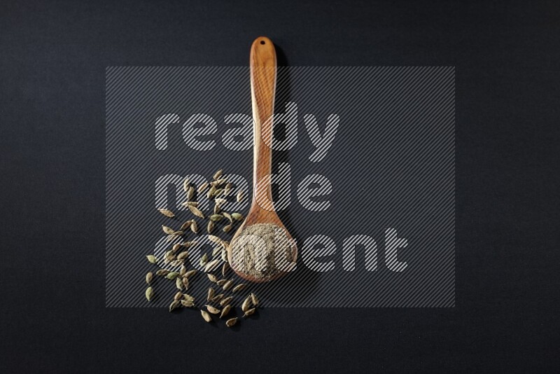 A wooden ladle full of cardamom powder and cardamom seeds beside it on black flooring