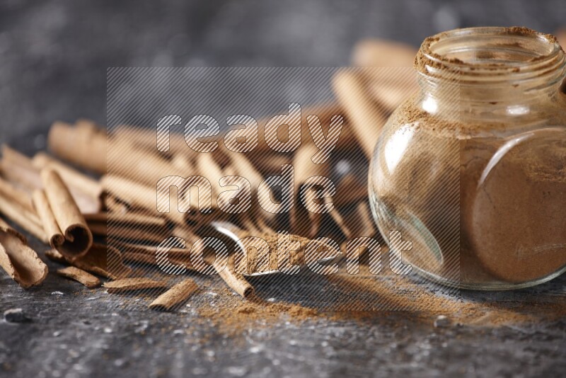 Herbal glass jar and a metal spoon full of cinnamon powder surrounded by cinnamon sticks on textured black background