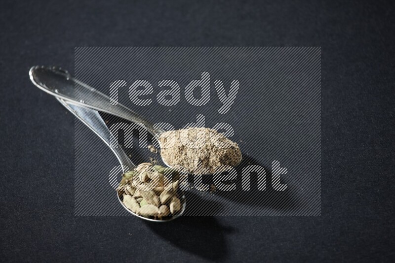 2 Metal spoons full of cardamom powder and cardamom seeds on black flooring