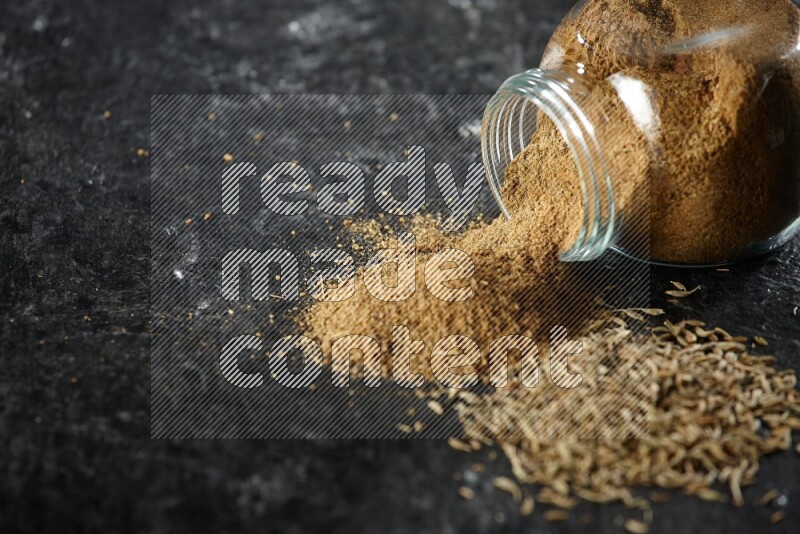 A flipped glass spice jar full of cumin powder with spilled powder and cumin seeds on a textured black flooring