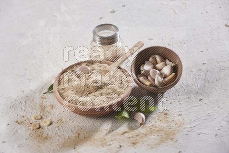 A wooden bowl with a spoon in it and glass spice jar, all full of garlic powder and a wooden bowl full of garlic cloves on a white flooring