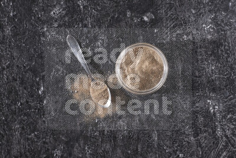 A glass jar full of black pepper powder and a metal spoon full of powder on a textured black flooring