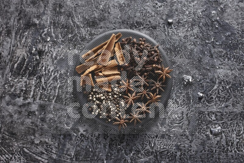 Cinnamon sticks, cloves, star anise and black and white peppers on a black plate on textured black background